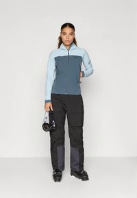 Donna con giacca a zip blu e grigia e pantaloni da sci neri in stile cargo tiene in mano degli occhiali da sci neri, in piedi contro uno sfondo bianco uniforme.