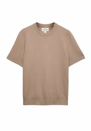 Giovane uomo che indossa un maglione beige a maniche corte, pantaloni color crema, scarpe marroni, tiene la tracolla di uno zaino marrone con una mano, guardando di lato.