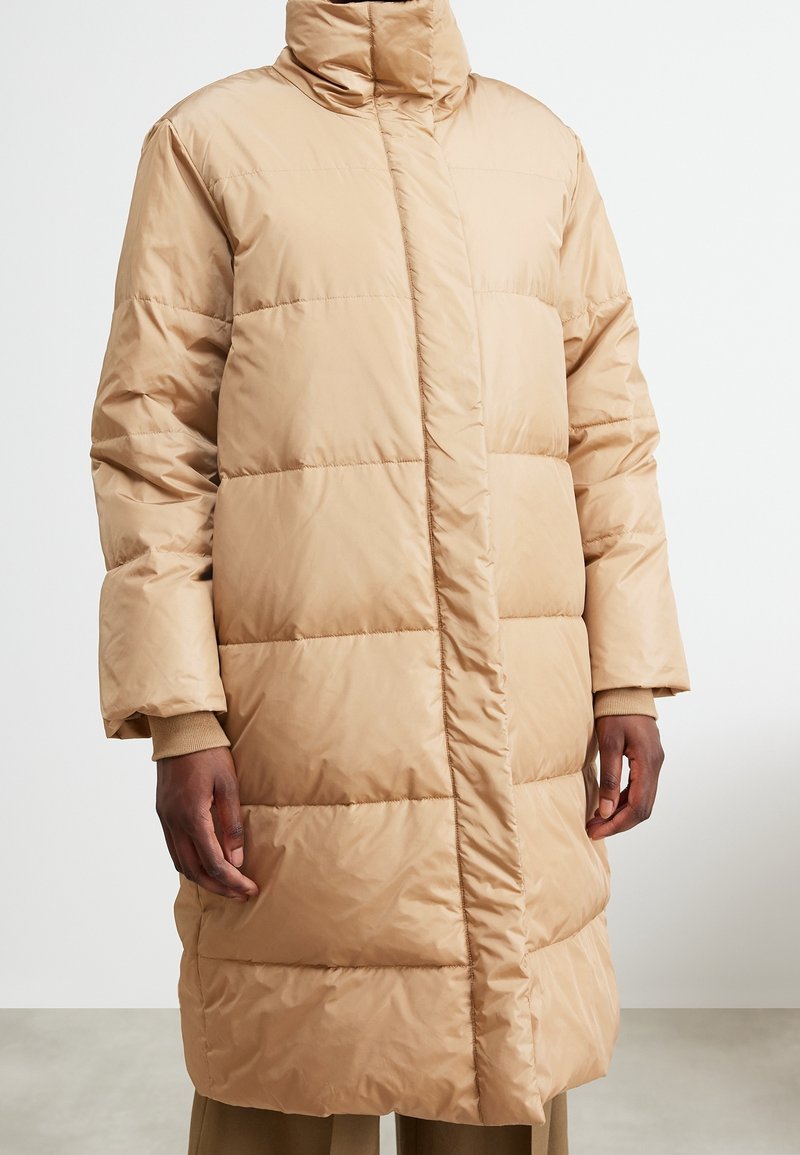 Long manteau beige matelassé avec col montant et fermeture avant dissimulée, porté sur un pantalon assorti et un pull à poignets côtelés.