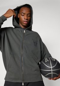 Felpa grigia con zip e logo di un toro, cappuccio con coulisse e tasche frontali. Il modello tiene un pallone da basket, mettendo in risalto uno stile atletico e casual.