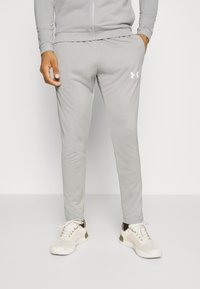Homme portant un pantalon de survêtement Under Armour gris clair et des chaussures de sport blanches, debout avec une main dans la poche devant un fond uni.