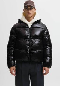 Uomo che indossa un giubbotto imbotitito nero lucido con foderina in shearling bianco, pantaloni scuri e berretto marrone, in piedi contro uno sfondo neutro.