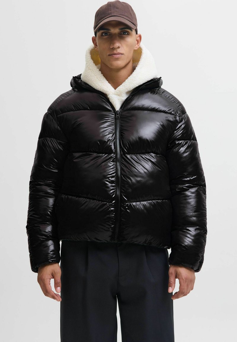 Uomo che indossa un giubbotto imbotitito nero lucido con foderina in shearling bianco, pantaloni scuri e berretto marrone, in piedi contro uno sfondo neutro.
