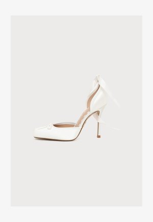 Zapato blanco de tacón alto con puntera puntiaguda, detalle de cinta en la parte delantera y correa en el tobillo atada en forma de lazo en la parte trasera.