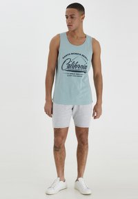 Lys blå tank topp med svart tekstgrafikk, kombinert med grå shorts. Modellen har på seg hvite sneakers og har en synlig tatovering på benet.