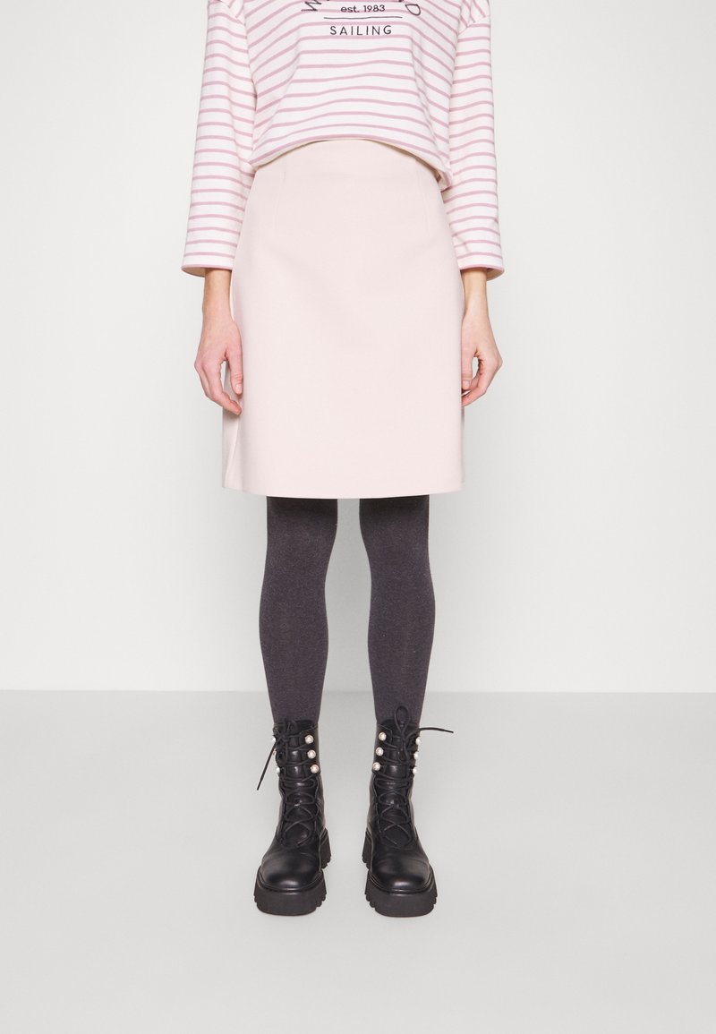 Femme portant une jupe rose pâle longueur genoux, un chemisier rayé à manches longues, des collants gris foncé et des bottes noires à lacets avec des détails en perles.