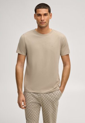 Mann steht mit den Händen in den Taschen und trägt ein beiges kurzärmliges Hemd sowie beige gemusterte Hosen vor einem einfarbigen Hintergrund.