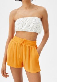 Top senza maniche in pizzo bianco con ritagli floreali, abbinato a pantaloni corti arancioni brillanti con vita elastica e tasche laterali.