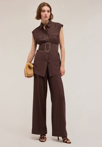 Salopette marrone senza maniche con cintura e bottoni, pantaloni a gamba larga e due tasche sul petto. Gli accessori includono una borsa a tracolla strutturata.