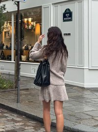 Lichtbeige gebreide trui gecombineerd met een bijpassende uitlopende rok. Zwarte leren handtas met een gouden kettinghengsel. Achtergrond van een straat met cobblestones.