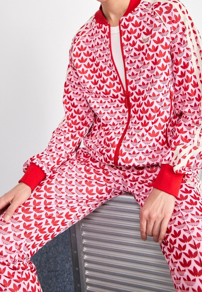 Röd och rosa sportdräkt med blommönster, bestående av en zip-up jacka och elastiska muddar, tillsammans med matchande byxor och vita detaljer.