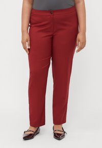 Vörös, slim-fit nadrág sima textúrával, egy gombos záródással és testreszabott kialakítással. Fényes bordó balerinacipővel viselve.