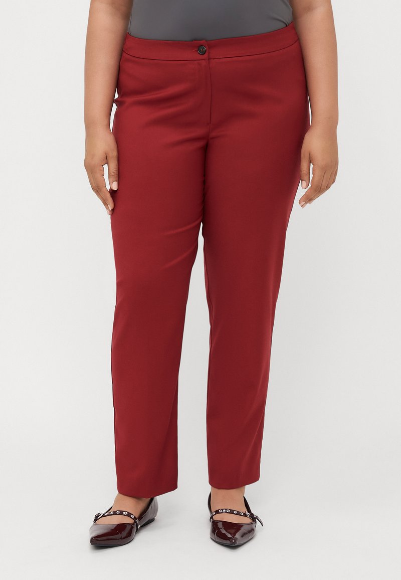 Vörös, slim-fit nadrág sima textúrával, egy gombos záródással és testreszabott kialakítással. Fényes bordó balerinacipővel viselve.