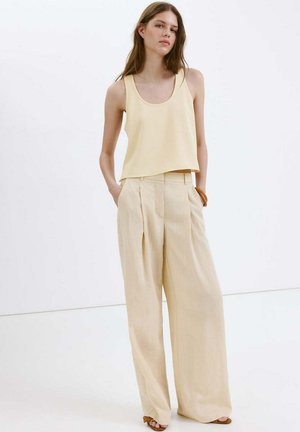 Femme vêtue d'un haut sans manches jaune pâle et d'un pantalon beige à jambes larges, debout les mains dans les poches, devant un fond blanc.