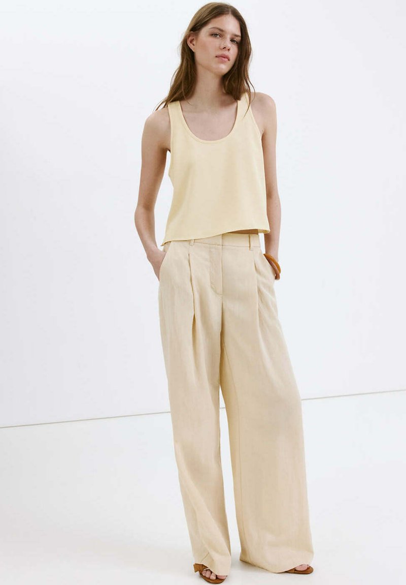 Femme vêtue d'un haut sans manches jaune pâle et d'un pantalon beige à jambes larges, debout les mains dans les poches, devant un fond blanc.