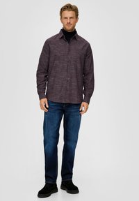 Langärmliges, knöpfbares Hemd in dunklem Lila mit einem strukturierten Muster, kombiniert mit blauen Jeans und schwarzen Schuhen, stehend vor einem neutralen Hintergrund.