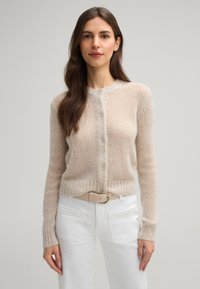 Helle beige, gestrickte cardigan mit Knopfleiste, runder Halslinie, langen Ärmeln und gerippten Bündchen. Kombiniert mit weißen Hosen und einem beigen Gürtel.