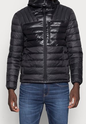 Veste matelassée noire Calvin Klein à capuche avec fermeture éclair devant et poche poitrine zippée, portée avec un jean bleu.