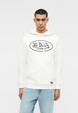 Mees seisab näoga otse vaataja poole, kandes valget kapuutsiga pusa musta "Von Dutch California" logoga ja helesiniseid teksaseid, taust on lihtne.