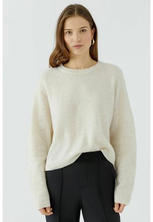 Femme aux cheveux bruns mi-longs portant un pull crème et un pantalon noir, debout devant un fond clair uni.