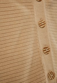 Bézs színű kötött felső, texturált bordákkal, a fronton nagy, mintás gombokkal. A közeli nézet kiemeli a szövet és a gomb részleteit.
