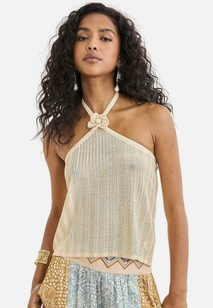 Mujer con cabello rizado que lleva un top halter beige con detalle de flores, falda estampada, pendientes colgantes y pulsera, posando frente a un fondo blanco.