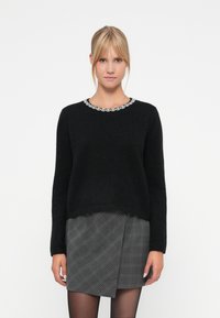 Black sweater with a round neckline featuring a decorative silver trim, paired with a gray plaid skirt. Soft texture, relaxed fit.