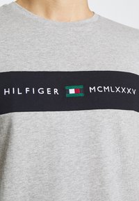 Camiseta de algodón gris con una banda horizontal negra. Texto bordado "HILFIGER MCMLXXXV" y un logo colorido en el centro.