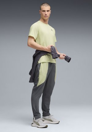 Chemise à manches courtes vert clair, pantalon gris foncé zippé avec des accents vert clair, pull gris noué autour de la taille et chaussures de course grises.