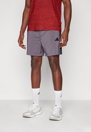 Pantalones cortos deportivos grises con tres rayas negras, combinados con una camiseta roja texturizada y calcetas blancas. Zapatillas blancas con detalles plateados completan el conjunto.