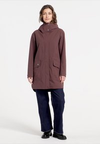 Waterdichte bruine jas met capuchon, drukknoopsluiten, twee voorzakken en een relaxed fit, gedragen over donkere jeans en laarzen.