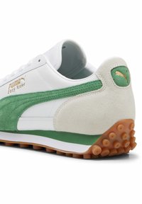 Zapatilla blanca con acentos de gamuza verde. Presenta un logotipo dorado, un collar acolchado y una suela de goma texturizada con picos salientes.