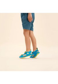 Sandali in tessuto blu con ritagli, suole gialle e cinturini regolabili. Abbinati a pantaloni corti, il design enfatizza comfort e traspirabilità.