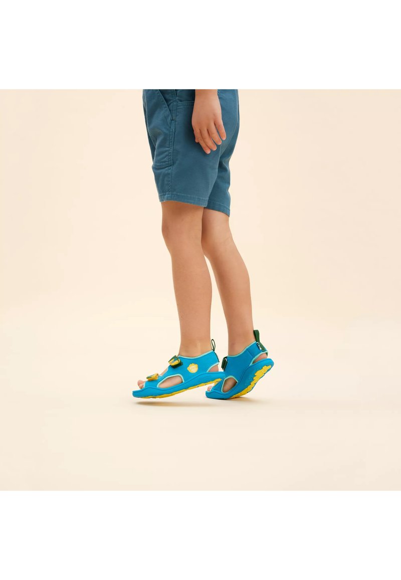 Sandali in tessuto blu con ritagli, suole gialle e cinturini regolabili. Abbinati a pantaloni corti, il design enfatizza comfort e traspirabilità.