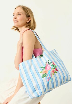 Young woman smiling, wearing pink top and white skirt, carrying blue and white striped tote bag with colorful parrot and floral design on shoulder.