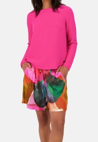 Fuchsia Langarmbluse kombiniert mit bunten Druckshorts, die abstrakte Formen in Pink, Grün, Orange und Braun aufweisen.