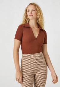 Brauner gerippter Kurzarm-Body mit einem tiefen V-Ausschnitt. Kombiniert mit hoch taillierten, gemusterten Hosen in einem geometrischen Design aus Braun und Creme.