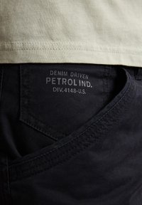 Czarne dżinsy z bawełnianego twillu z widoczną kieszenią, z szarym logo "DENIM DRIVEN PETROL IND.", teksturowana powierzchnia i staranne szwy.