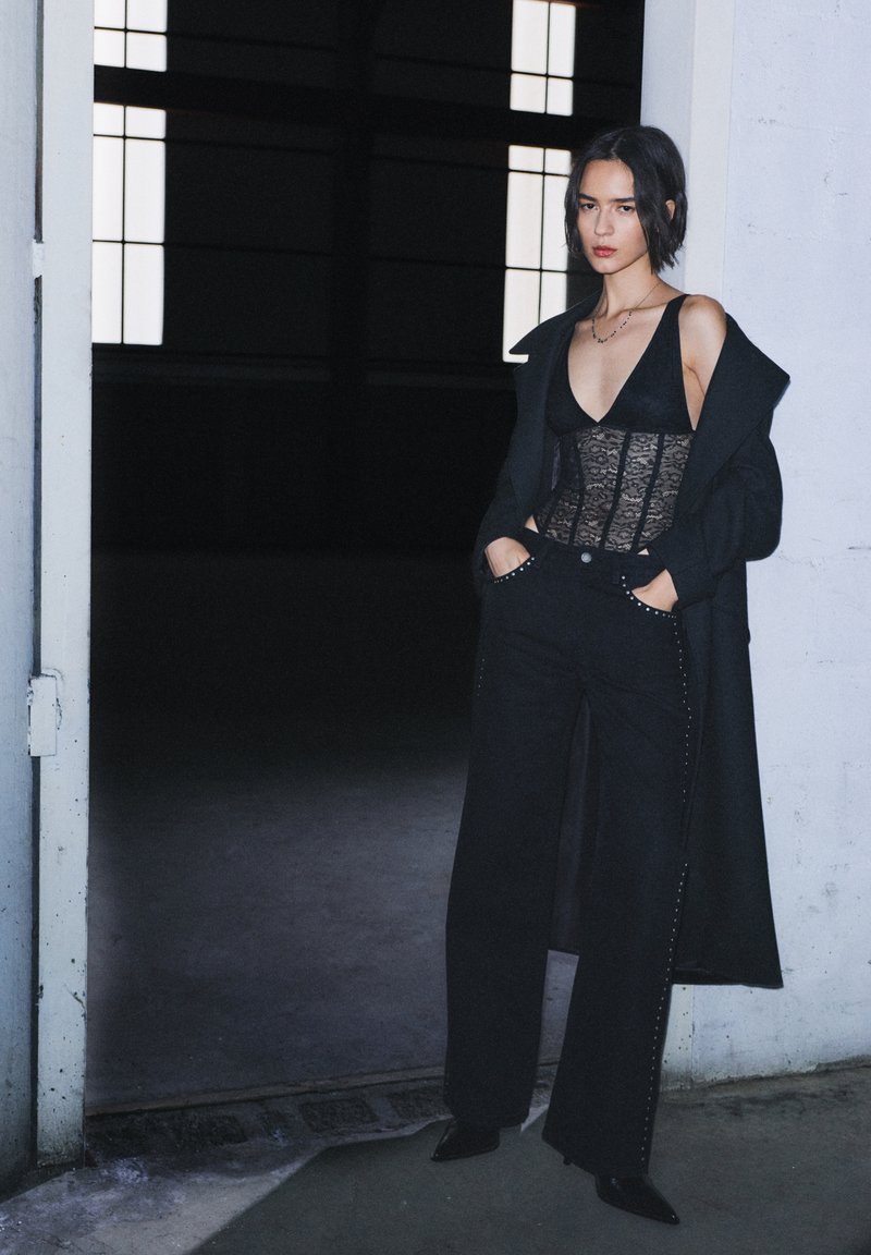 Fekete csipke body hosszú fekete kabát alatt, széles szárú fekete farmerrel, amely díszes szegecsekkel van ellátva, sötét háttér előtt.