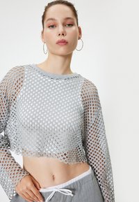 Lichtgrijze crop top met een mesh ontwerp met ronde openingen, lange mouwen en een ronde halslijn, gelaagd over een witte top.