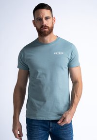 Petrolblauwe katoenen t-shirt met korte mouwen, ronde hals en een kleine witte logoafbeelding op de linkerborst. Gemaakt in combinatie met donkere spijkerbroek.