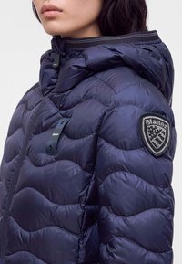 Femme portant une doudoune matelassée bleu marine avec une capuche et un patch logo Blauer sur la manche supérieure.