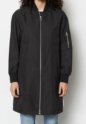Mittellange schwarze Bomberjacke mit silbernem Frontreißverschluss, Reißverschlusstasche am linken Ärmel, gerippten Bündchen, getragen über hellblauer Jeans.