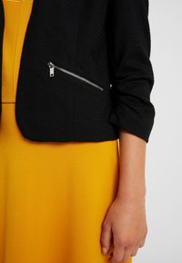 Black textured cropped jacket with a zippered pocket. Worn over a bright yellow dress, showcasing a smooth fabric finish.