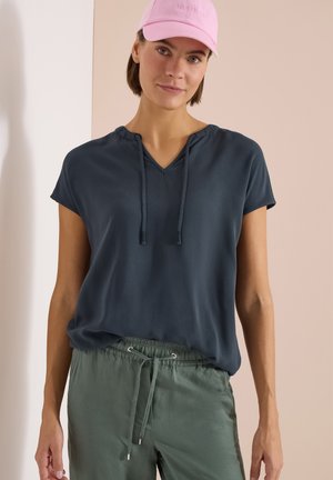 Femme portant une casquette de baseball rose, un haut à manches courtes bleu foncé avec des liens et un pantalon vert à cordon de serrage, debout face à un fond neutre.