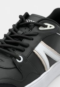 Baskets en cuir noir avec des accents blancs et beiges, bout arrondi et lacets noirs. Présente des perforations pour la ventilation et une semelle en caoutchouc blanc.