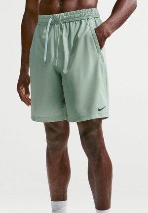 Mand iført lysegrønne Nike træningsshorts med snøre i taljen og hånd i lommen, står mod en ensfarvet baggrund.