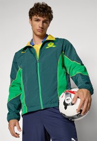 Grønn og gul sportjakke med glidelåser, med kontrasterende sidepaneler og et emblem. Holder en fotball med svart og hvit design.