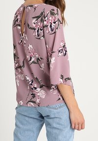 Blommig blus i mjuk mauve-nyans med nyckelhålsrygg. Har trekvartslånga ärmar och en lös passform. Kontrasterande mörkgröna och rosa blommor.