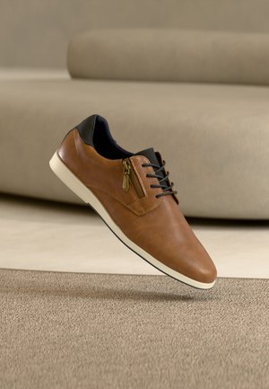 Chaussure décontractée en cuir marron avec col noir, fermeture éclair latérale, lacets noirs et semelle blanche, posée sur un tapis beige avec un arrière-plan neutre.